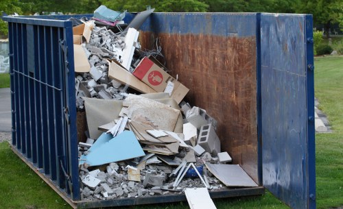 A large blue industrial skip filled with a variety of construction and building waste, including broken concrete slabs, scattered plasterboard, and discarded wooden and metal debris, situated outdoors on a grassy area with a partially open side panel revealing the clutter inside. The skip's surface appears weathered with some rust visible on the interior, and it is positioned on a paved or gravel surface. In the background, there are trees and greenery suggesting a suburban or park-like setting, closely related to rubbish removal services in the Covent Garden area. The overall scene reflects a typical waste disposal situation, with debris spilling over the sides and the surrounding environment indicating an active rubbish collection or clearing operation supported by companies such as Commercial Waste Covent Garden.