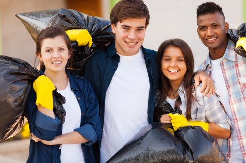 Four young adults standing outdoors in an urban environment, each holding large black rubbish bags filled with waste. The group consists of two women and two men, all smiling and wearing casual clothing, with some wearing yellow work gloves. The rubbish bags appear to contain general waste and are slightly crumpled, with visible textures of plastic and black colour. The individuals are positioned close together, with a sidewalk and blurred city background behind them, suggesting a community clean-up or rubbish clearance activity. The scene reflects a focus on waste management and rubbish removal services available in Covent Garden, London. Commercial Waste Covent Garden’s service involves collecting and disposing of rubbish generated by households or businesses, emphasizing responsible waste handling in local areas. The natural lighting highlights the integrity of the waste bags and the camaraderie of the group, subtly indicating the importance of efficient rubbish removal in urban environments.