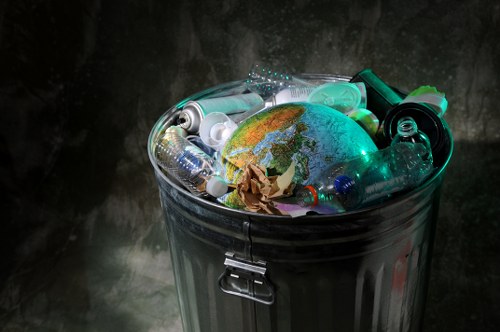 A metal rubbish bin with a closed lid, filled to the brim with various discarded plastic bottles, crumpled paper, a small globe depicting Earth's continents with a blue, green, and brown surface, and other miscellaneous waste materials. The bin is positioned against a dark, textured background, likely within an indoor or shaded outdoor setting. The plastic bottles are transparent and of different shapes and sizes, some with caps still attached. The crumpled paper appears brown and reused, contrasting with the smooth surface of the globe and the glossy finish of the plastic waste. The scene suggests a focus on waste disposal, emphasizing the need for proper rubbish removal services, such as those offered by Commercial Waste Covent Garden, in urban environments like central London. The lighting highlights the reflective surfaces of the bottles and the globe, creating a somber, observational tone relevant to environmental waste management.