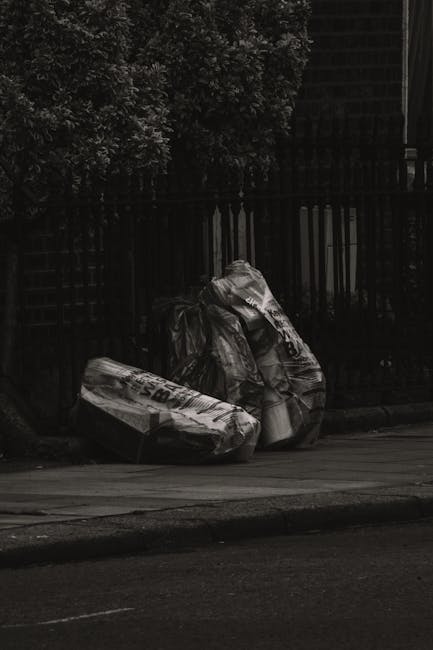 A narrow urban alleyway cluttered with large, black and white trash bags filled with rubbish, positioned in the foreground near a wheeled trolley or sack, with surrounding walls covered in graffiti an